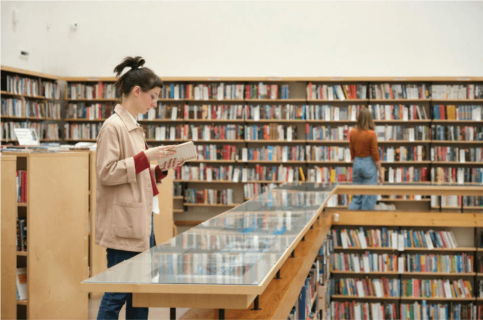 A girl stands reading a book in a library, surrounded by bookshelves