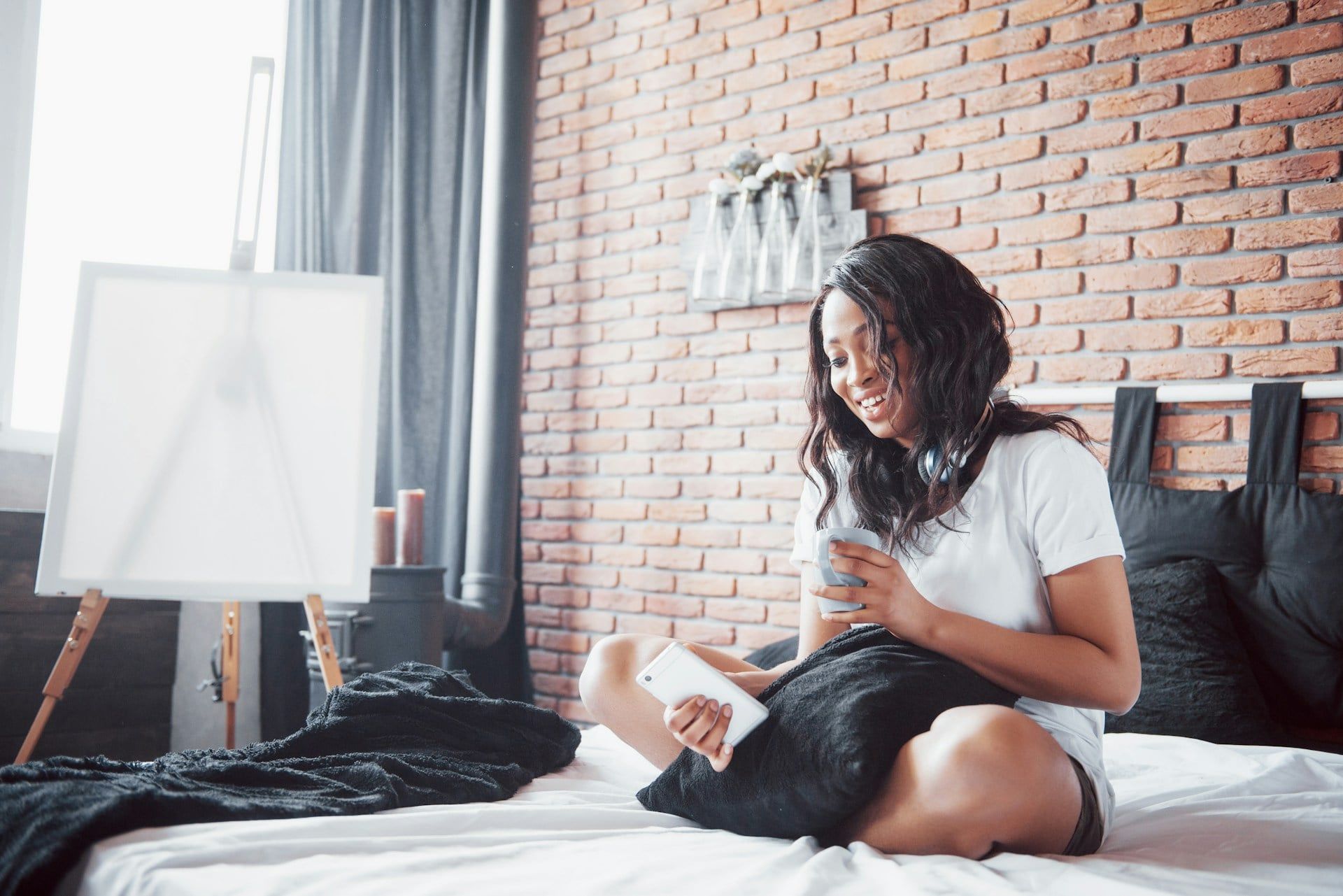 A woman sits cross-legged on her bed using her smartphone, smiling with a drink