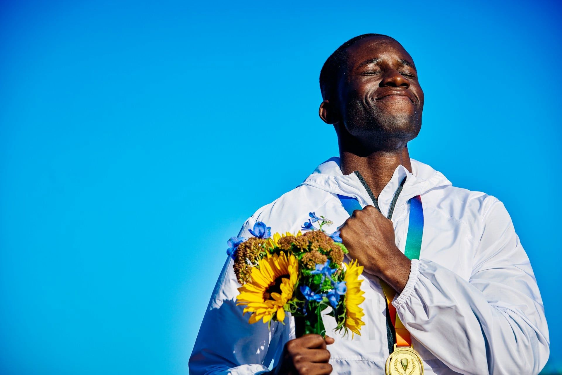 A man smiles holding flowers, a medal, and a hand to his chest after winning a competition