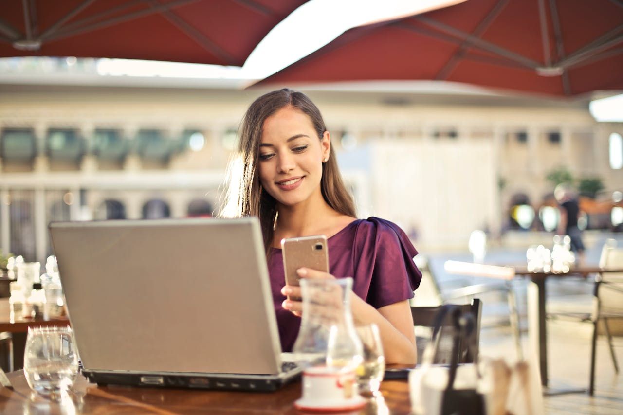 A woman sits working on her laptop while answering messages on her smartphone