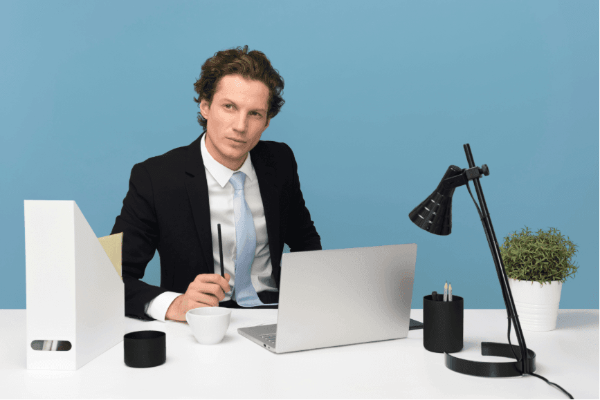 A man seated at a desk, working on a laptop, set against a plain blue background with a cluttered surface filled with office supplies.