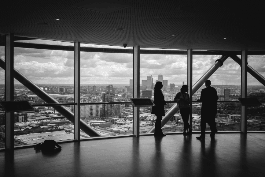 A grey-toned image of people standing by large glass panes with a view of the downtown skyline.