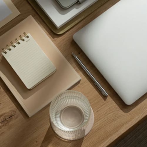 Carnet, stylo, ordinateur et verre d'eau posé sur une table, l'ensemble est dans les ton bois et gris clair