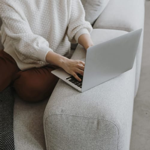 Femme assise sur un canapé gris vêtue d'un pull blanc qui tape sur un ordinateur portable