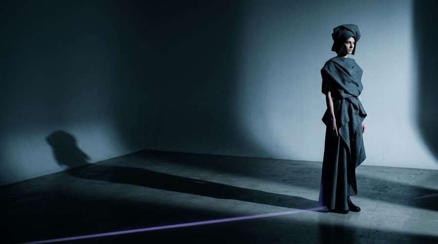 A model in a sculptural charcoal garment in an empty cold-blue studio