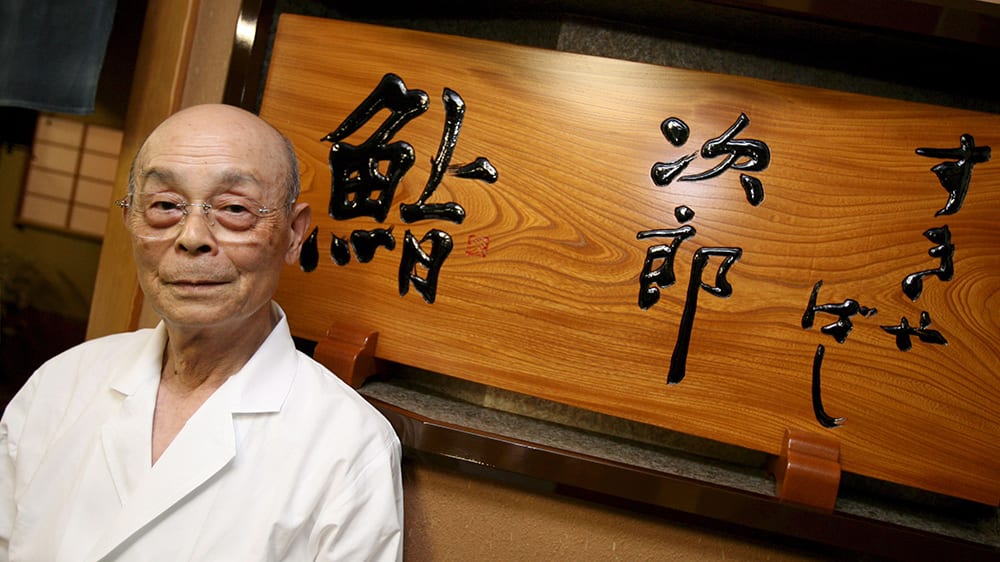 <strong>Jiro Ono</strong>, a famed Japenese chef, is considered the greatest living sushi master. He is the subject of the film, <em>Jiro Dreams of Sushi</em>, which provides a glimpse into Sukiyabashi Jiro, his Michelin three-star restaurant where his singular obsession with sushi is on display. (Photo Credit: <a href="https://robbreport.com/food-drink/dining/sukiyabashi-jiro-stripped-of-three-michelin-stars-2883698/">Robb Report</a>)