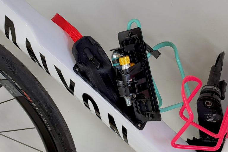 The storage hatch under the bottle cages, open: the black LOAD Tool Pack with its red pull strap tucked into the down tube, and the CO2 cartridge and inflator clipped inside the hatch cover