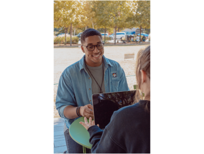 Two people sit facing each other at a table on a sidewalk. One has their back to the viewer with an open laptop in front of them