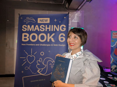 Cover designer Chiara Aliotta holding Smashing Book 6 in her hand in front of the Smashing books stand at SmashingConf NY