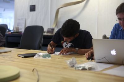 Two people sit next to each other at a long table. One person is using a laptop and mouse, the other person is sketching something on a sheet of paper.