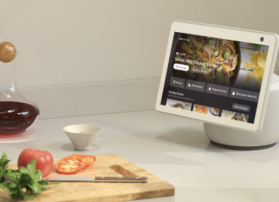 In the picture, Echo Show stands on a kitchen table next to a chopping board with some food on it.