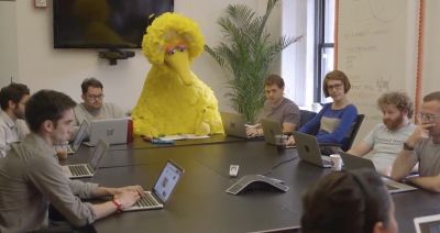 A meme photo showing Big Bird from Sesame Street sitting in a room full of designers with laptops