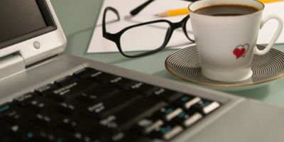 An of a computer, a cup filled with coffee with glasses, pencil, and paper in the background.