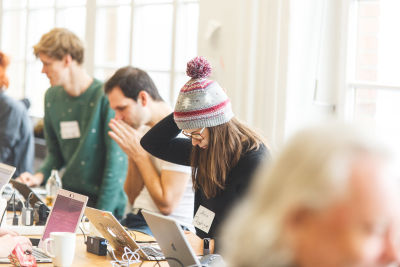 A photo of Ana Rodrigues at a previous IndieWebCamp working on their blog with other participants in the background