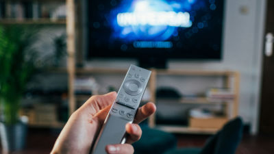 A hand holds a modern TV remote, aimed at a television screen. The blurred background emphasizes the viewing distance, illustrating the '10-foot experience,' a key aspect of TV interaction distinct from phones and computers.