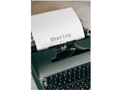 A manual typewriter is displayed. A white sheet of paper is in the typewriter with the word “Sharing” typed out on the page