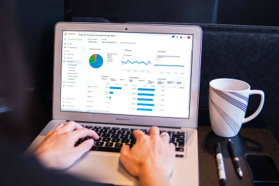 A photo displaying a laptop on a desk. Two hands rest on the laptop keyboard. The laptop screen displays various charts reflecting an analytics dashboard.