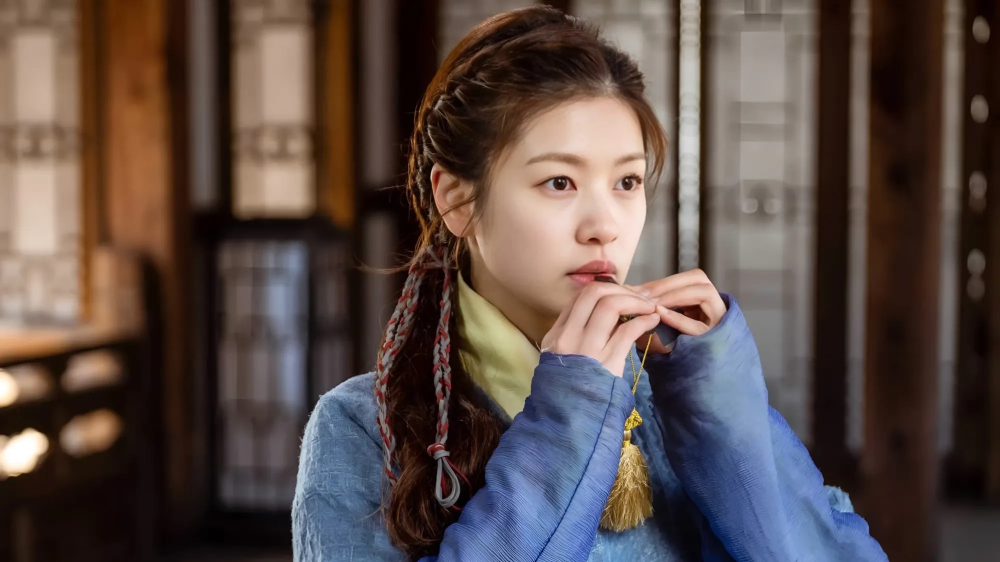 Mu-deok in a blue hanbok with braided hair, holding a small whistle to her lips inside a traditional wooden hall
