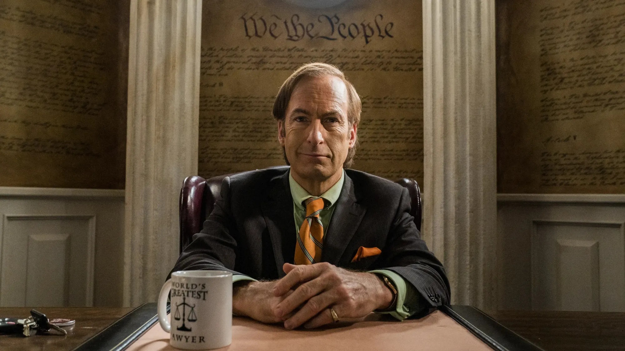 Saul Goodman sitting behind his desk in a dark suit and orange tie, hands folded, a World's Greatest Lawyer mug beside him and a painted Constitution backdrop behind
