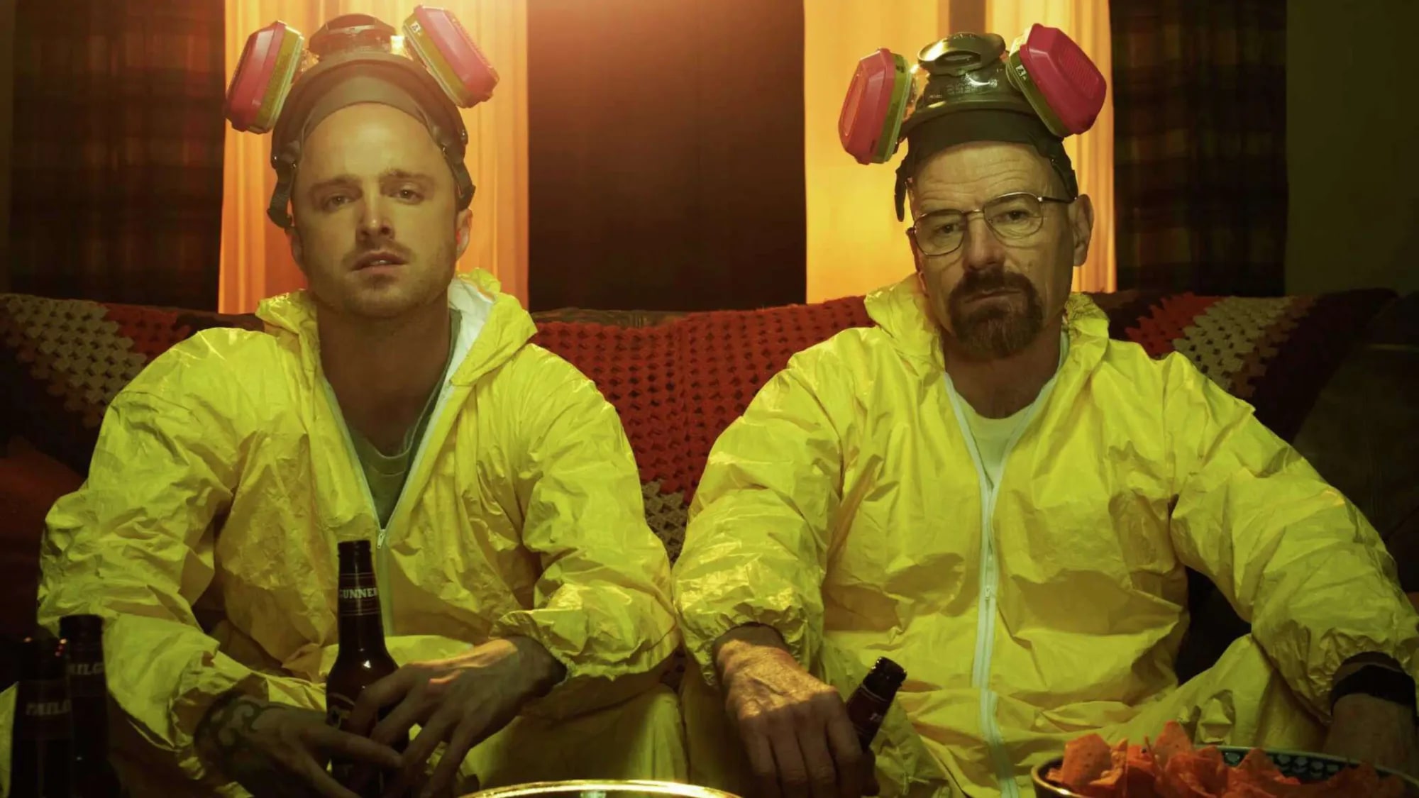 Jesse and Walt slumped on a couch in yellow hazmat suits with respirators pushed up onto their heads, beer bottles in hand, the room lit amber