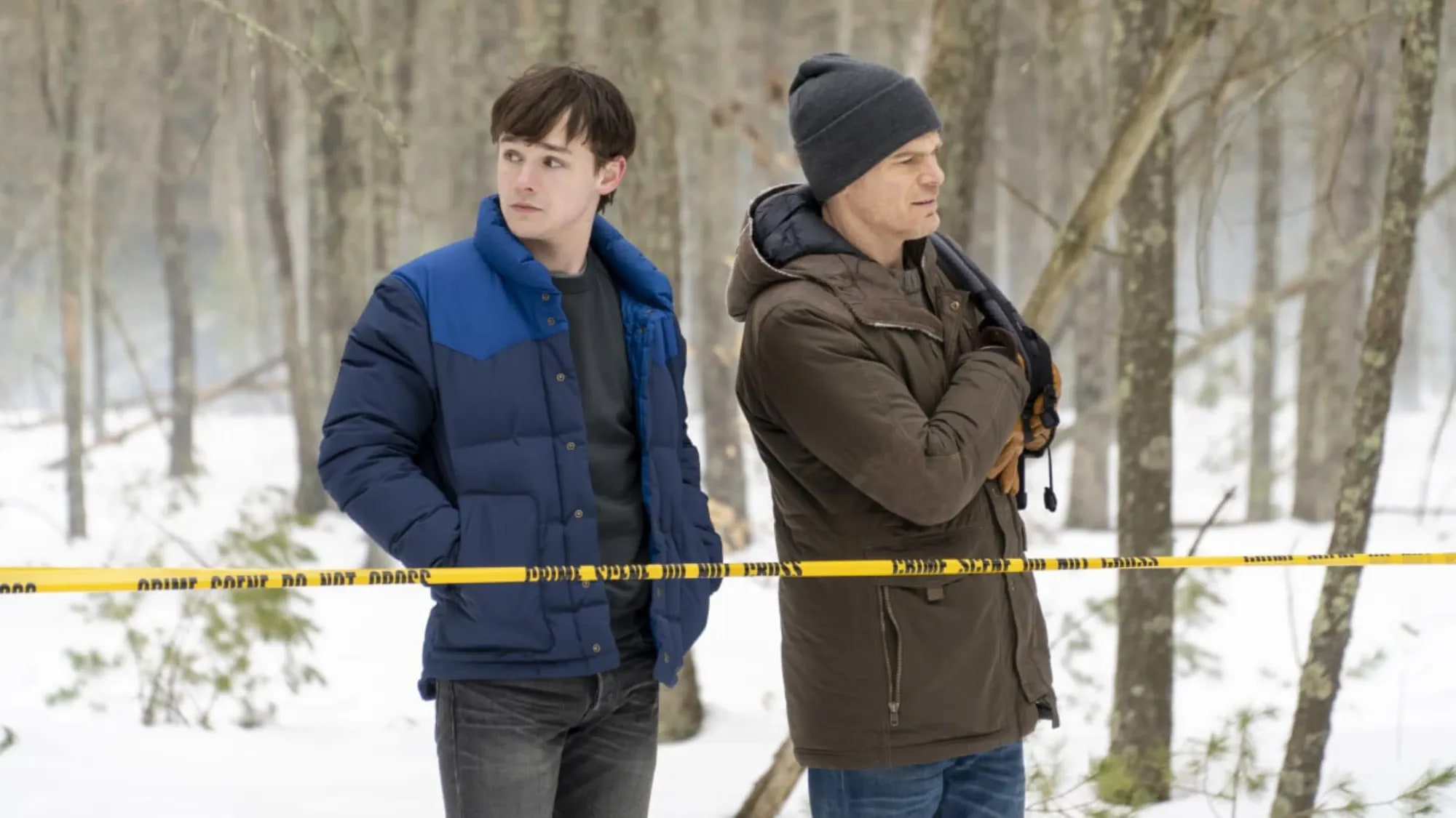 Harrison and Dexter standing behind yellow crime scene tape in snow-covered woods, both in winter coats, looking off to one side