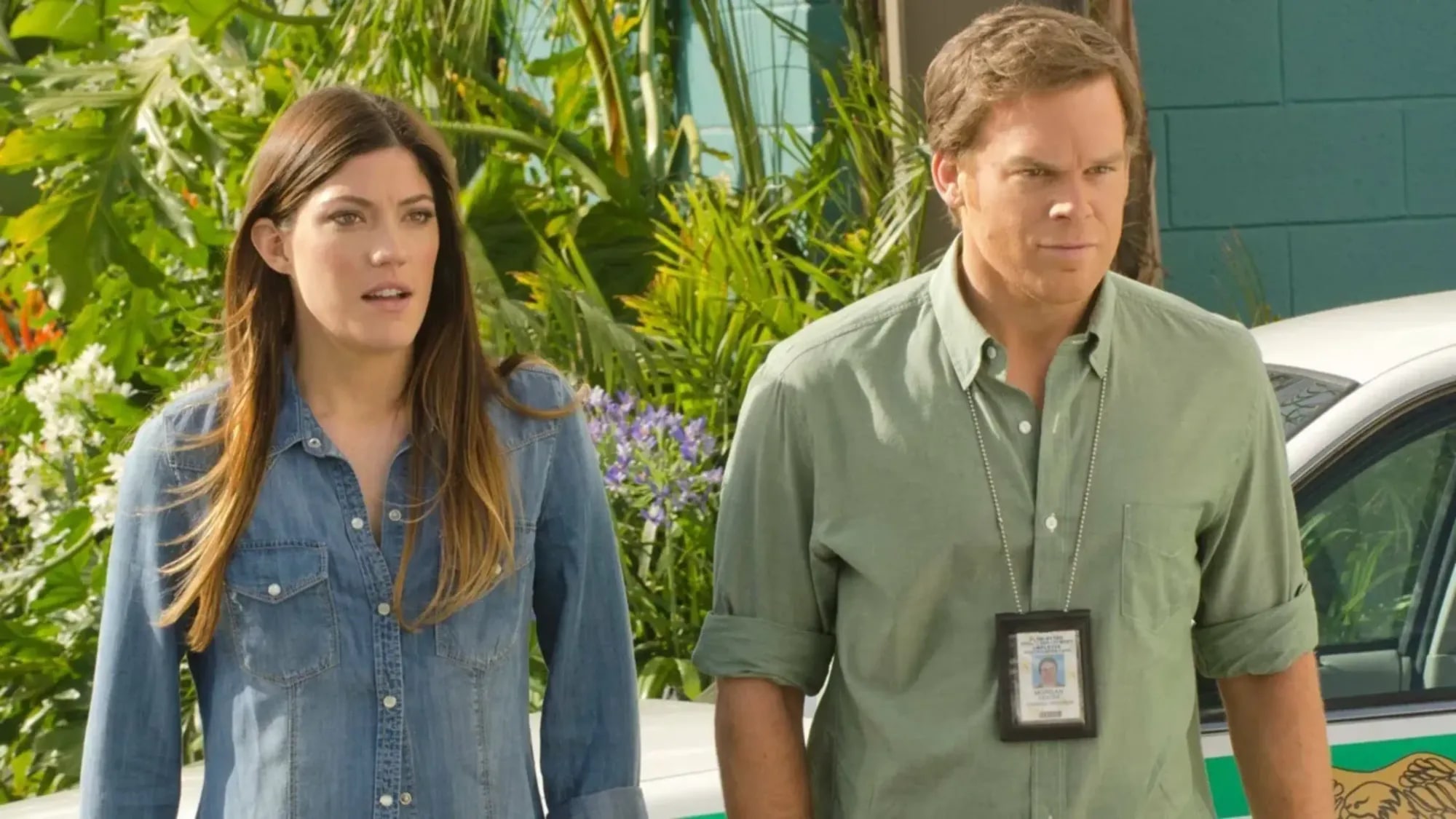 Debra and Dexter Morgan walking away from a squad car outside a Miami building, Dexter in a green shirt with his police ID on a lanyard