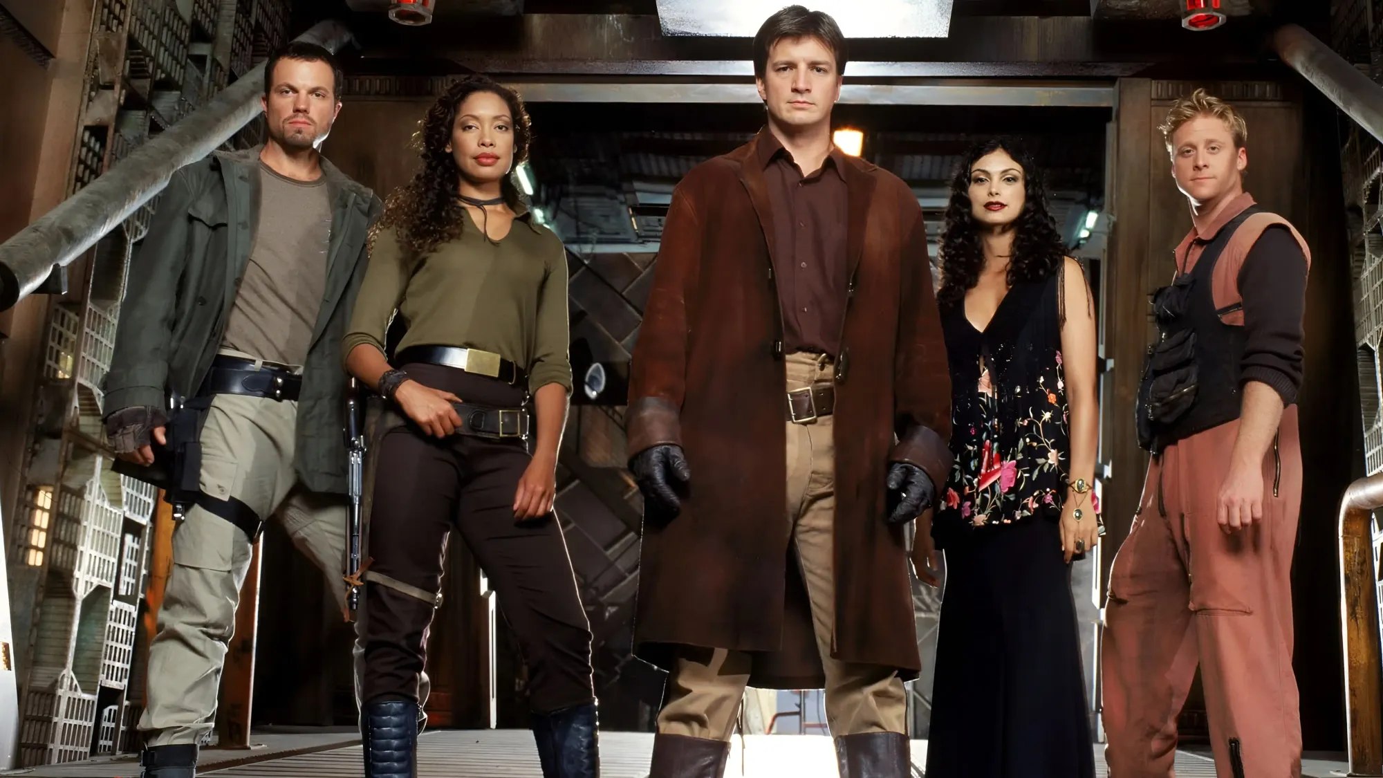 Five of the Serenity crew standing in a line in the ship's cargo bay, Mal at the centre in a long brown coat, the others armed and facing the camera