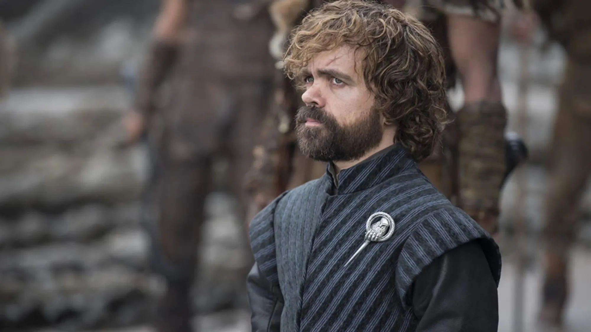 Tyrion Lannister wearing the Hand of the Queen pin, looking up at something out of frame, with armed guards blurred behind him