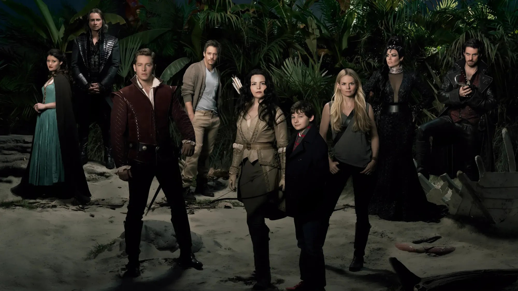 The main cast in fairy-tale and modern costume standing on a moonlit jungle beach, Snow White with a bow and Henry at the centre, Captain Hook and the Evil Queen to the right