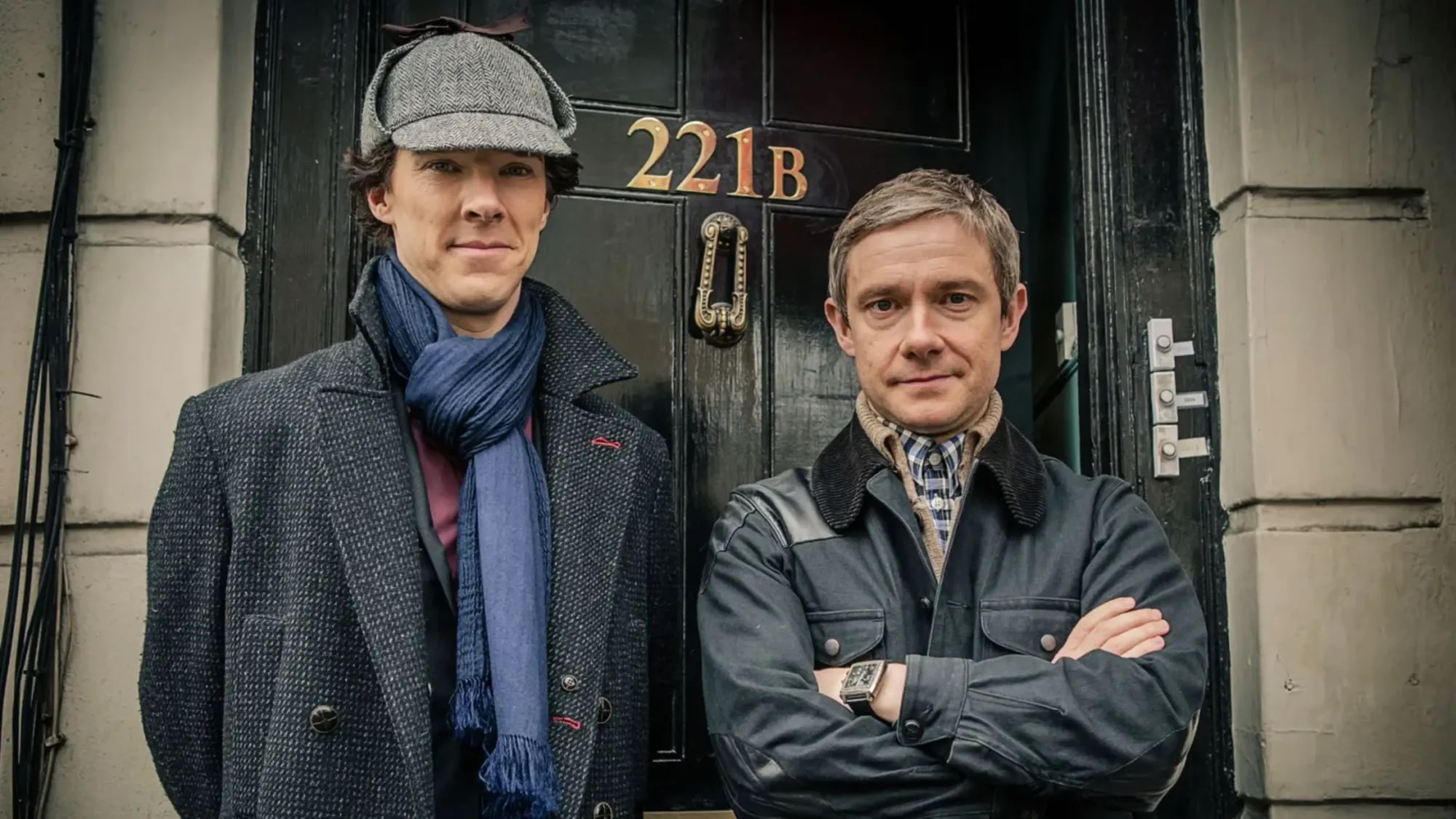 Sherlock in a deerstalker and blue scarf standing beside Watson, arms folded, in front of the black front door of 221B Baker Street