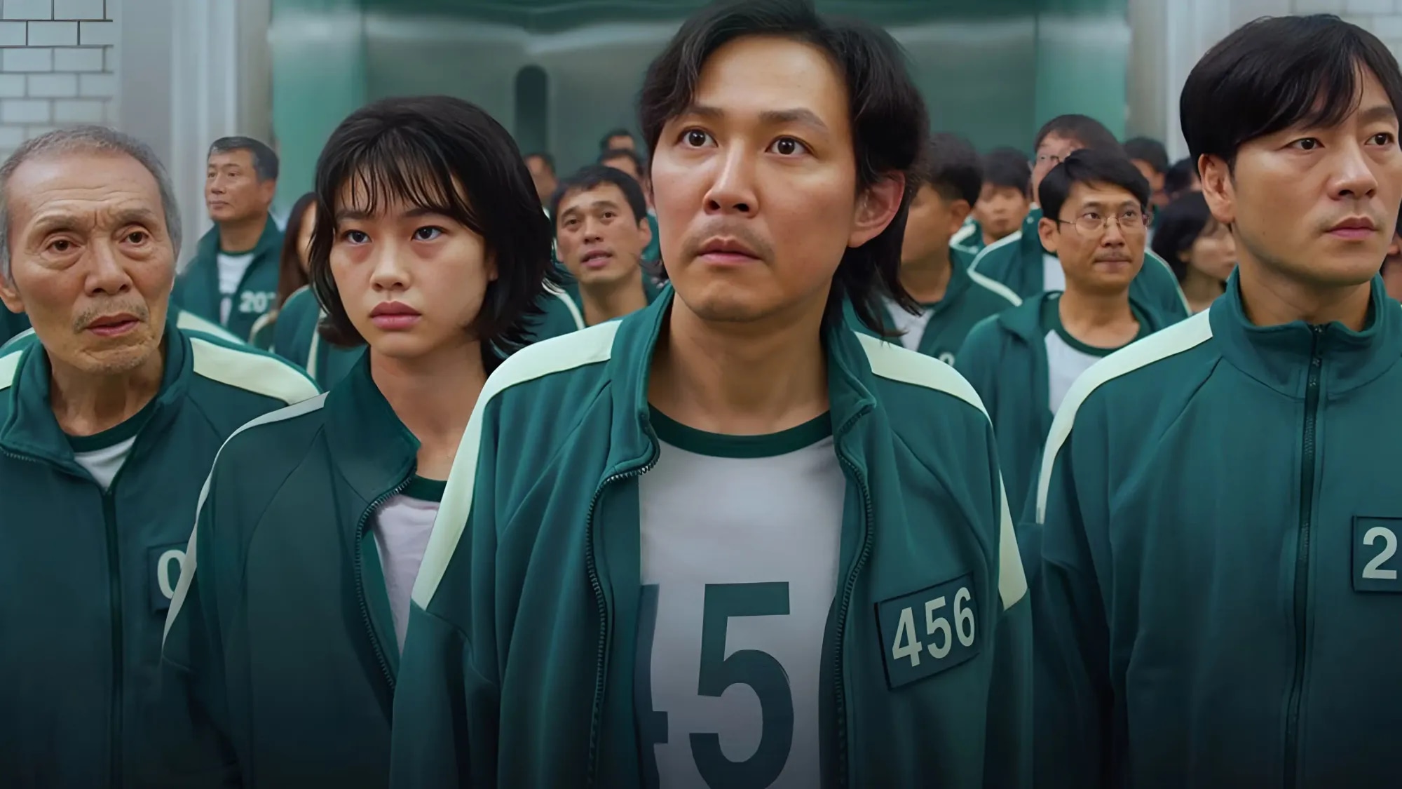 Gi-hun, player 456, standing in a crowd of players in green tracksuits and staring upwards in shock
