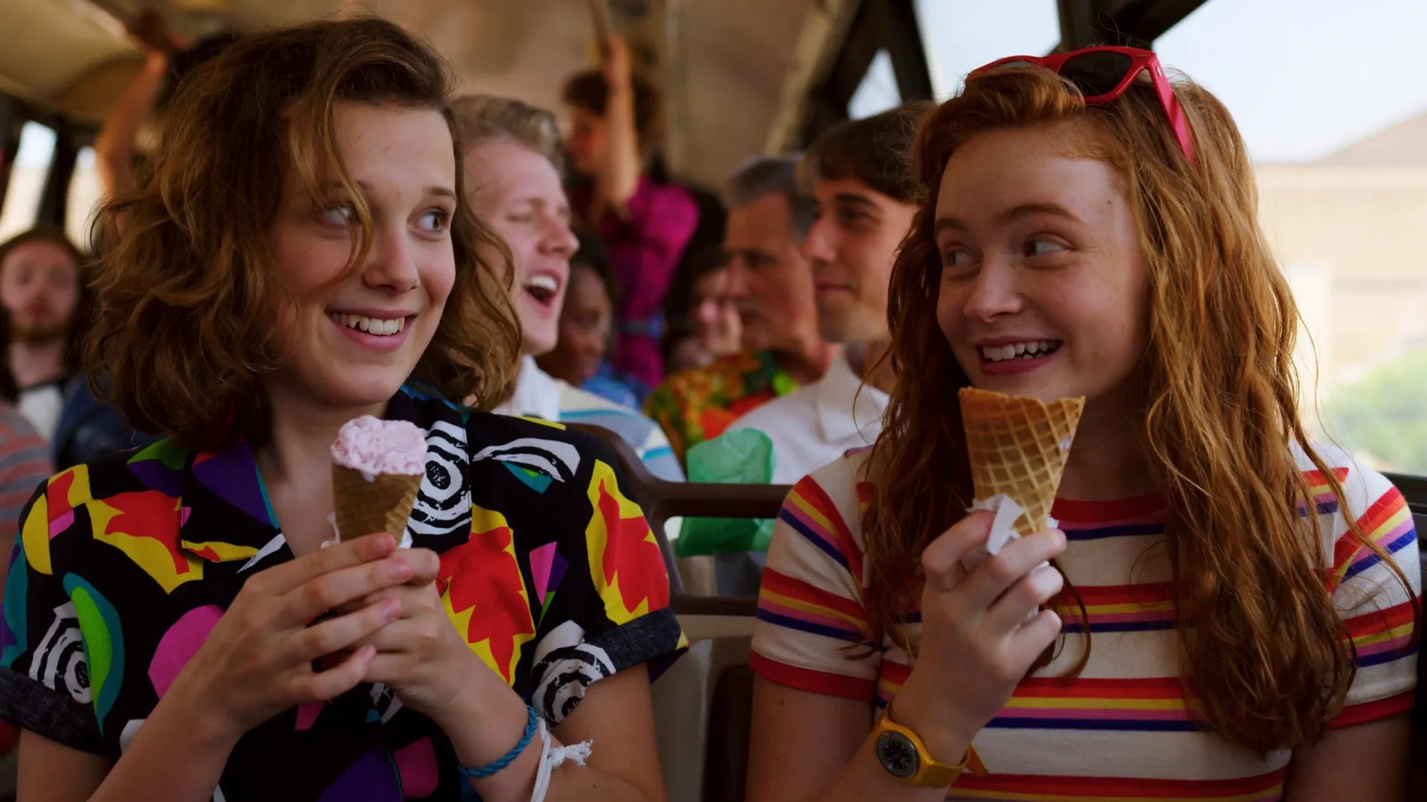 Eleven and Max sitting side by side on a crowded bus, each holding an ice cream cone and grinning at each other