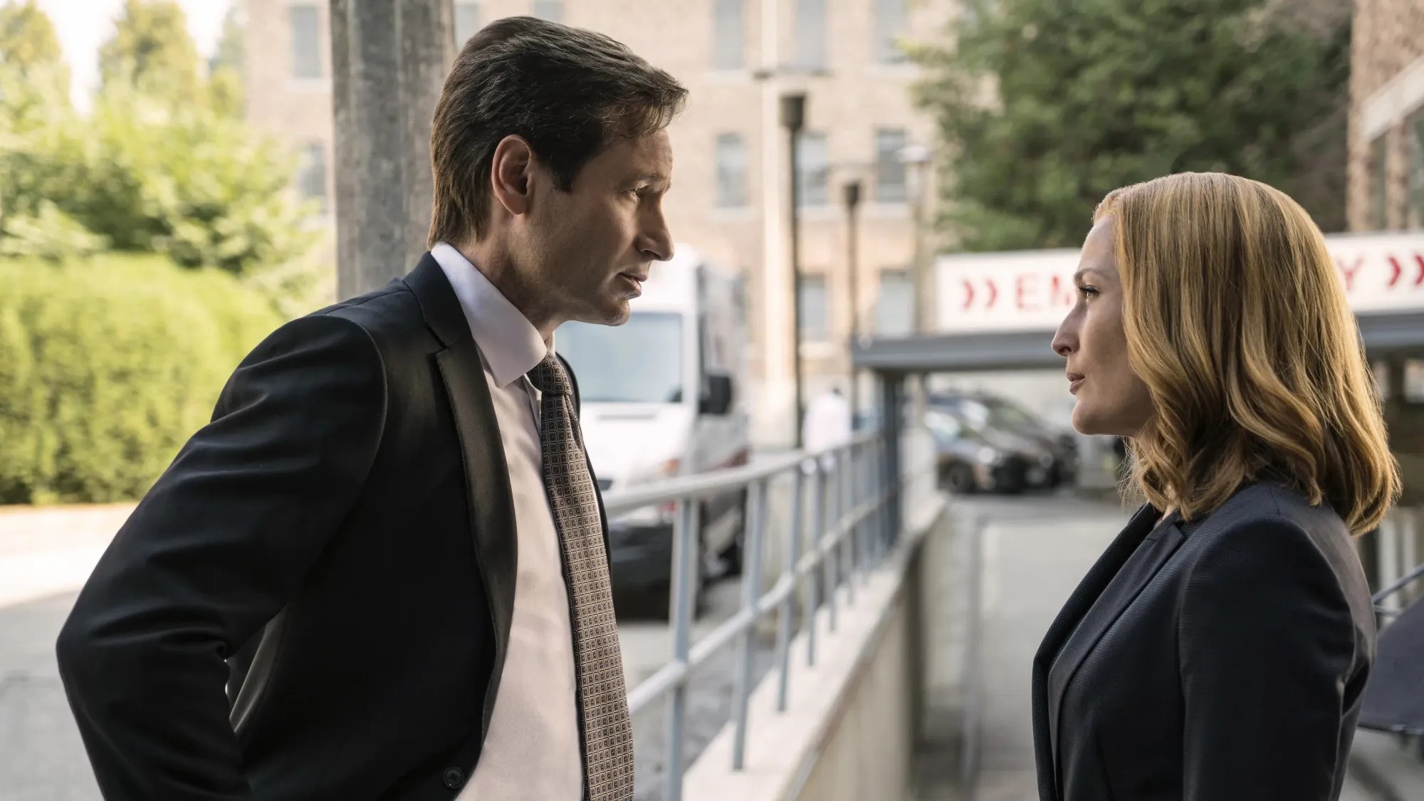 Mulder and Scully facing each other in dark suits outside a hospital emergency entrance, deep in conversation