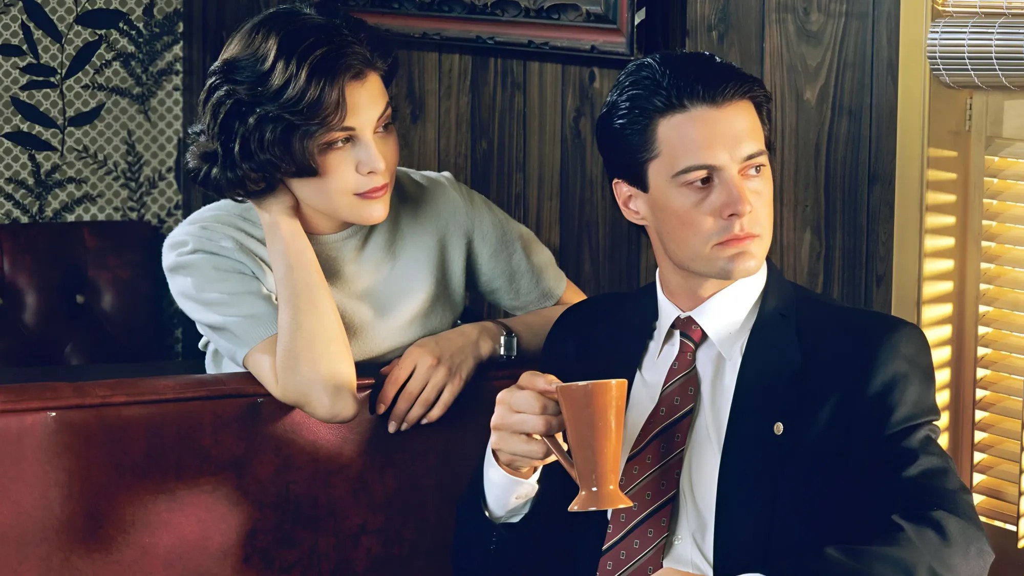 Audrey Horne leaning over the back of a diner booth, chin resting on her hand, watching Agent Cooper below her in a dark suit holding a mug of coffee