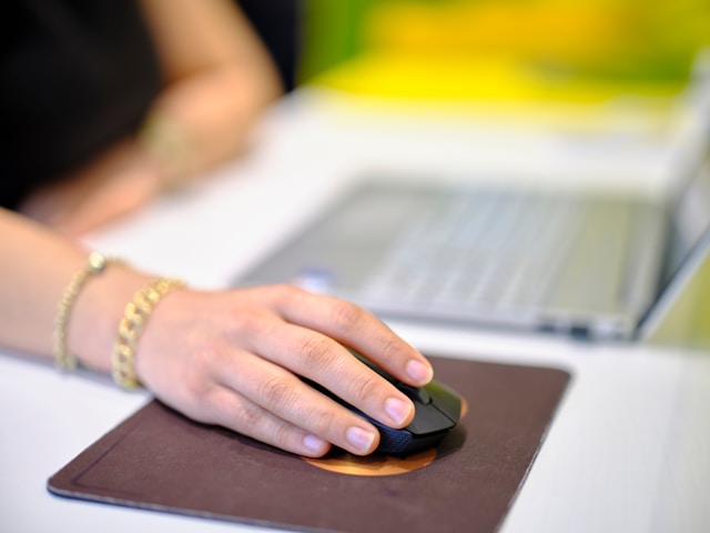 A person's hands using a computer mouse on a desk, with a blurred background of a laptop and other office supplies.