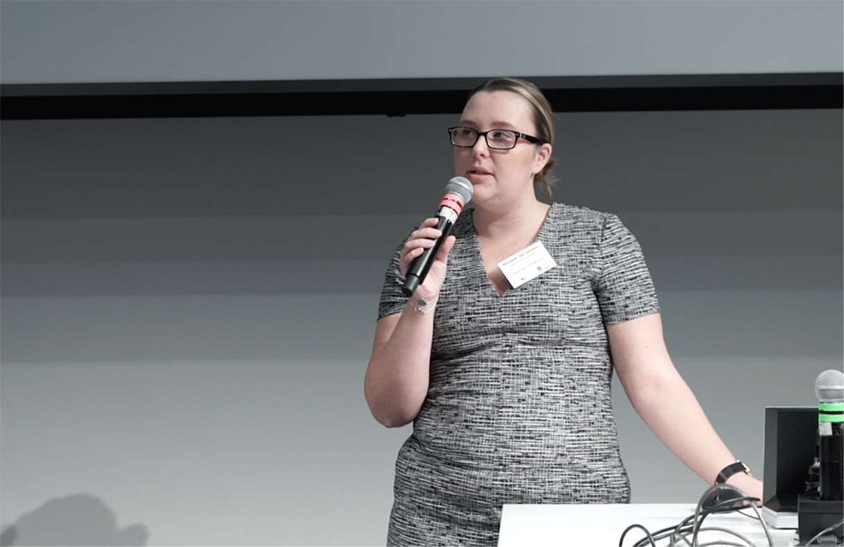 Monique standing at a lectern holding a microphone, speaking in a presentation setting. She is wearing a patterned dress and has a name badge clipped to her chest. A computer monitor and cables are visible on the desk beside her.
