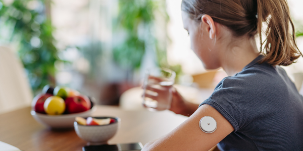 A person's arm with a fitness tracker on the wrist, holding a glass of water, with a bowl of fresh fruit in the background.