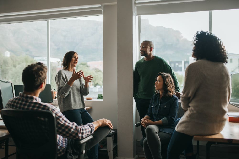 A group of people, both men and women, are gathered in a cozy room with large windows overlooking a scenic outdoor landscape.