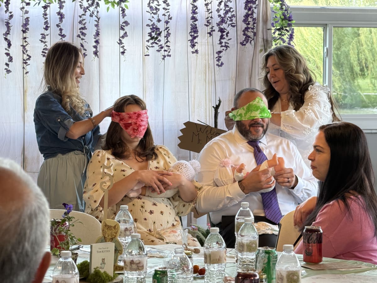 Bert and Vanessa sitting in chairs with baby dolls, trying to put diapers on them while blindfolded.