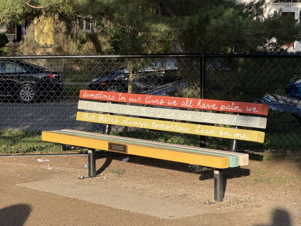 A rainbow painted park bench that reads: Sometimes in our lives we all have pain, we all have sorrow. But if we are wise, we know that there's always tomorrow. Lean on me.