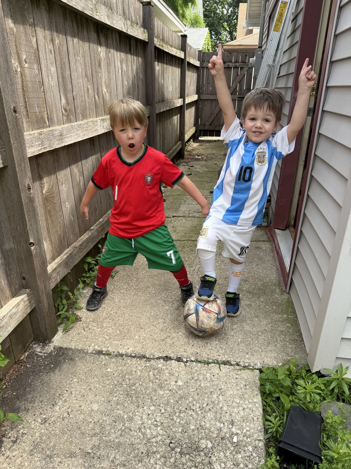 Ryan in a red Ronaldo Portugal kit and Owen in a white and blue Messi Argentina kit. Ryan is imitating Ronaldo’s signature celebration and Owen is stepping on a soccer ball with his arms raised victoriously in the air.