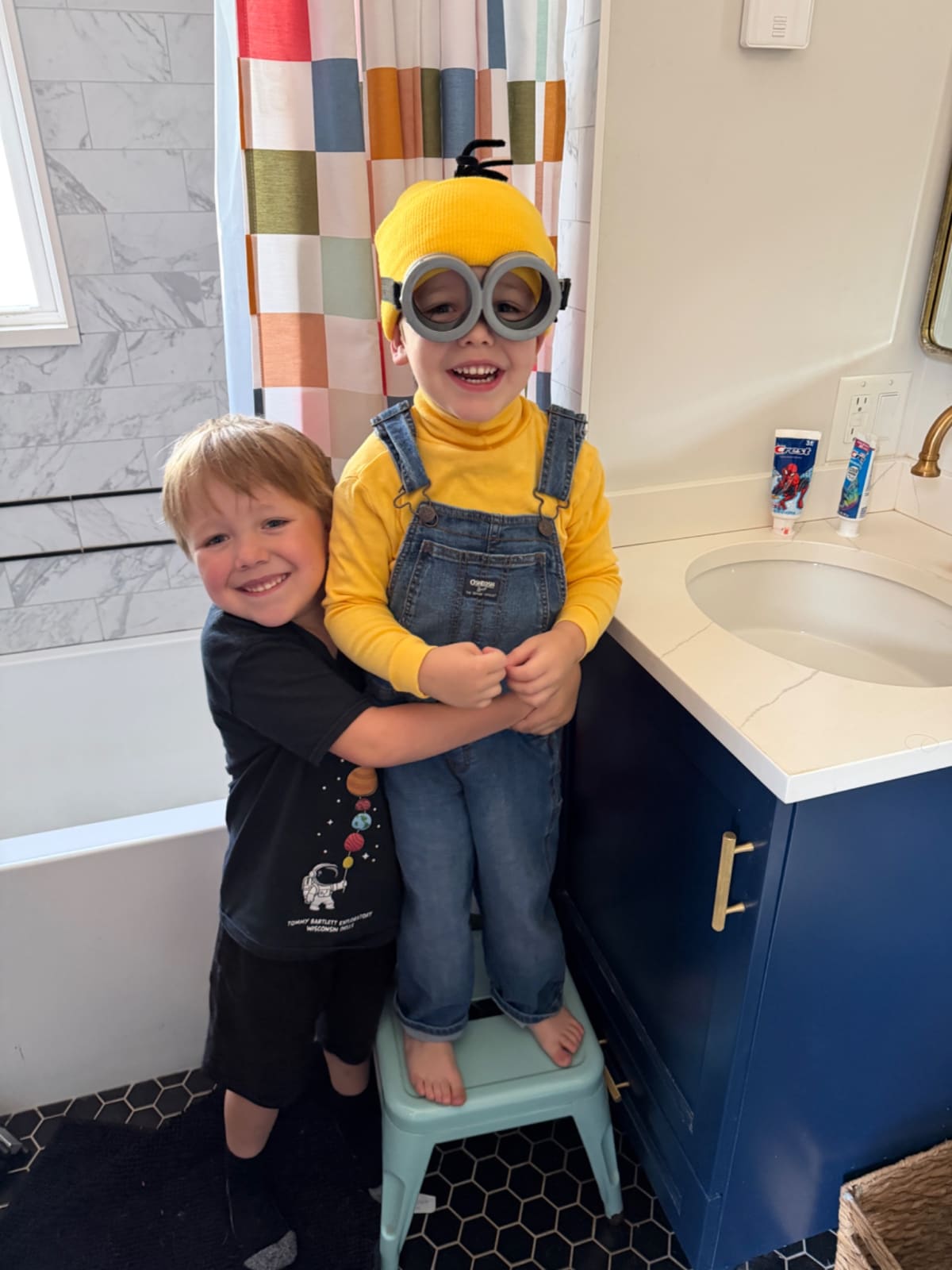 Owen standing on a stool next to the bathroom sink wearing a Minion costume. Ryan is standing next to him and hugging him.