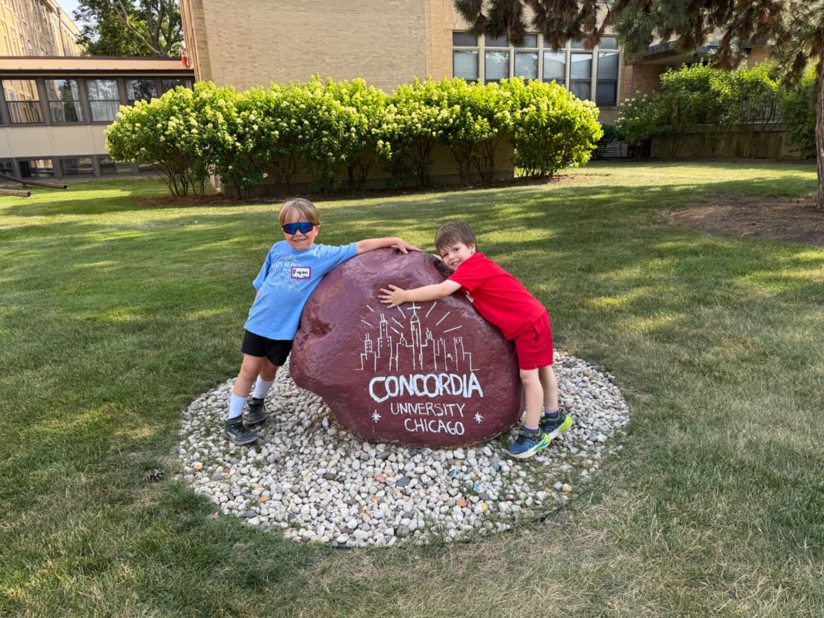 Ryan and Owen hugging a large, red rock with a cityscape and “Concordia University Chicago” painted on it.