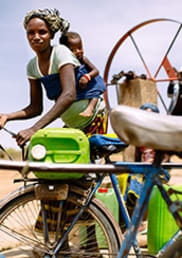 Woman with baby on her back untying a jerry can from a bicycle as she prepares to collect water. Two other people stand behind her at a pump.