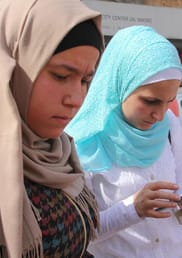 Two women map points on their phone. A woman on the left wears a beige hijab and the woman on the right wears a light blue hijab and looks at her phone.