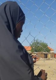 A girl with a headscarf and her back to the camera looks sadly at a wire fence. Blue skies and trees can be seen in the background.