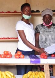 Julliene and her daughter stand to the right of their , Mauelle, at the market selling their goods at the market. They stand behind a market stall covered with tomatoes and bananas.