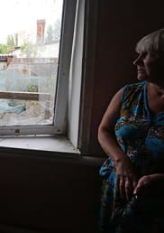 A woman with blond hair and a blue colourful dress sits to the right of a window. Buildings and trees can be seen in the background through the window.