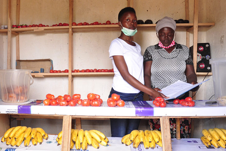 Julliene and her daughter stand to the right of their , Mauelle, at the market selling their goods at the market. They stand behind a market stall covered with tomatoes and bananas.