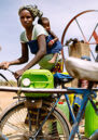 Woman with baby on her back untying a jerry can from a bicycle as she prepares to collect water. Two other people stand behind her at a pump.